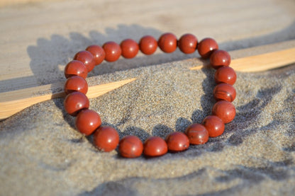 Bracelet Jaspe Rouge en pierre naturelle posé sur du sable à cheval sur un plateau en bois. perle de diamètre 7-8mm