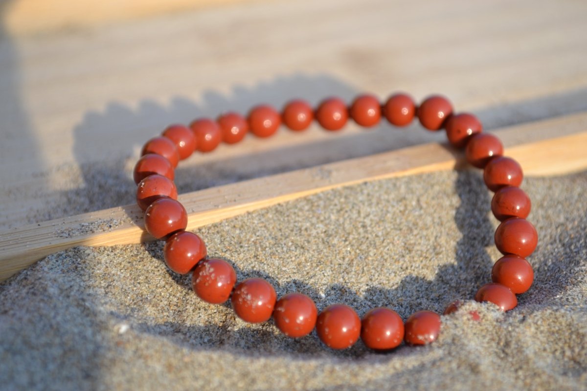 Bracelet Jaspe Rouge en pierre naturelle posé sur du sable à cheval sur un plateau en bois. perle de diamètre 5-6mm